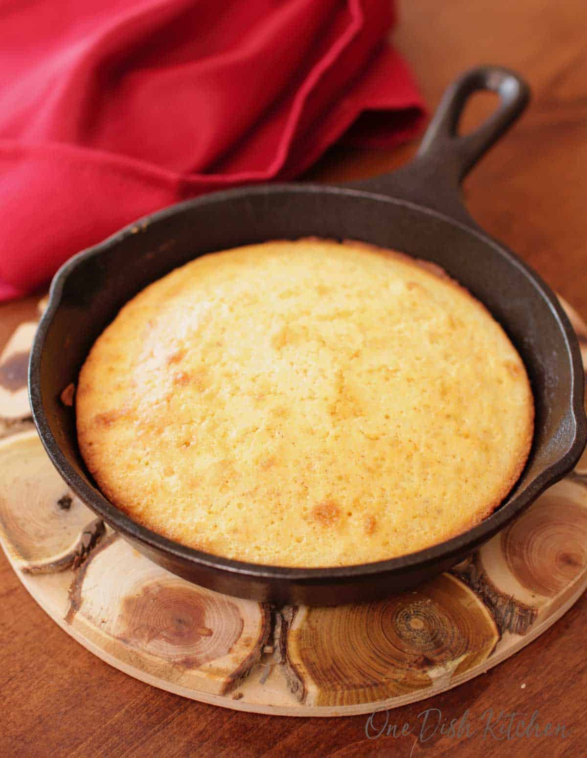 Cornbread in a small cast iron skillet next to a red napkin on a wooden trivet.