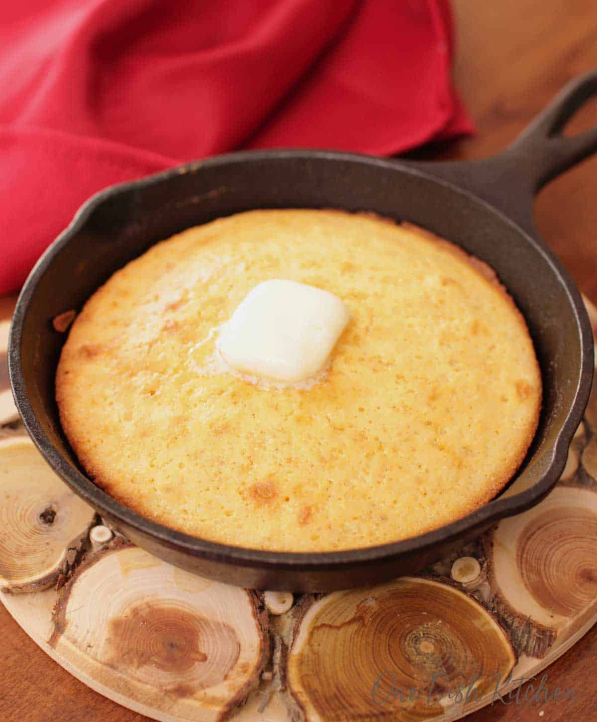 Butter melting on cornbread in a cast iron skillet on a wooden trivet next to a red cloth napkin.