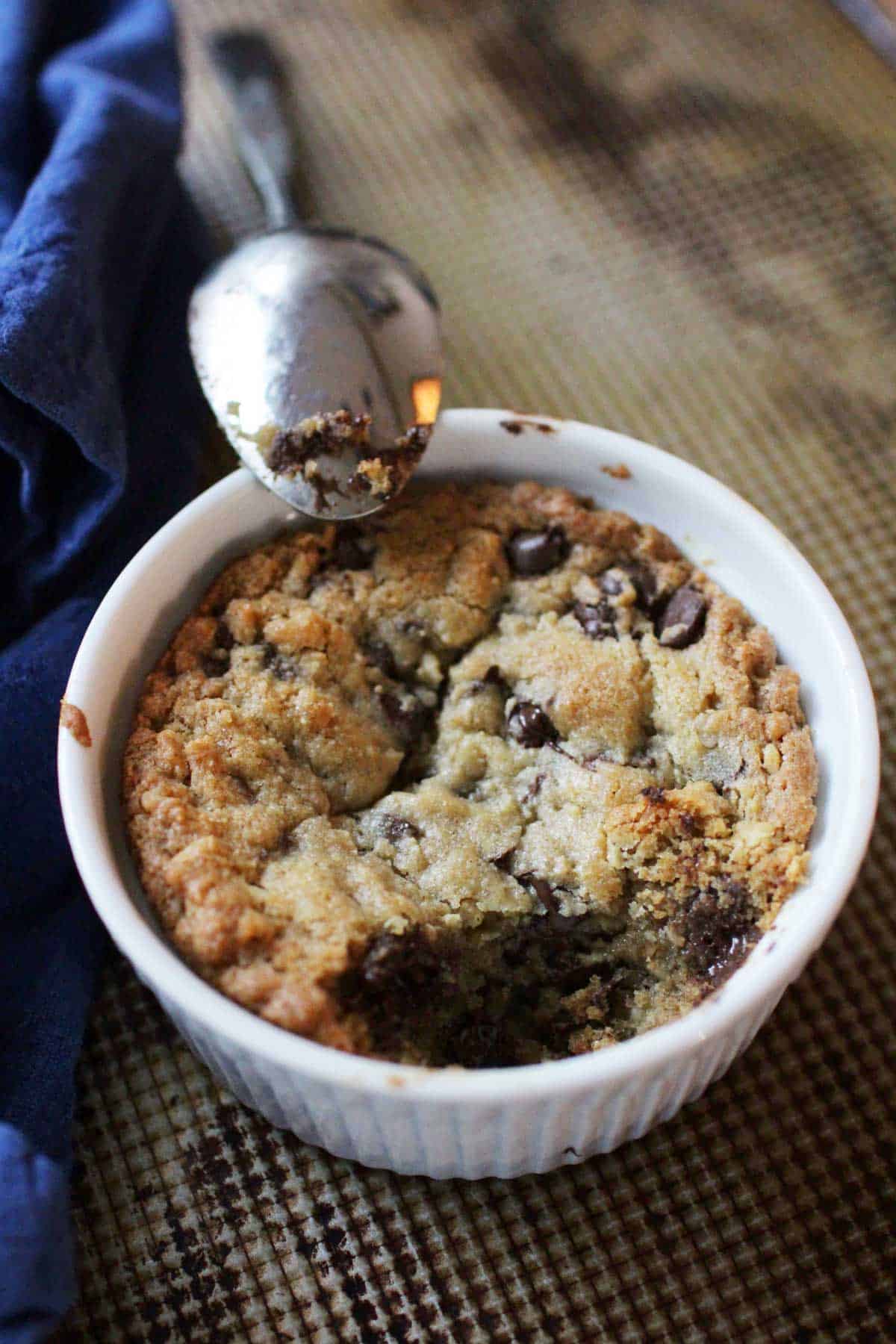 A spoon resting on the edge of a ramekin filled with a deep dish cookie made with chocolate chips.