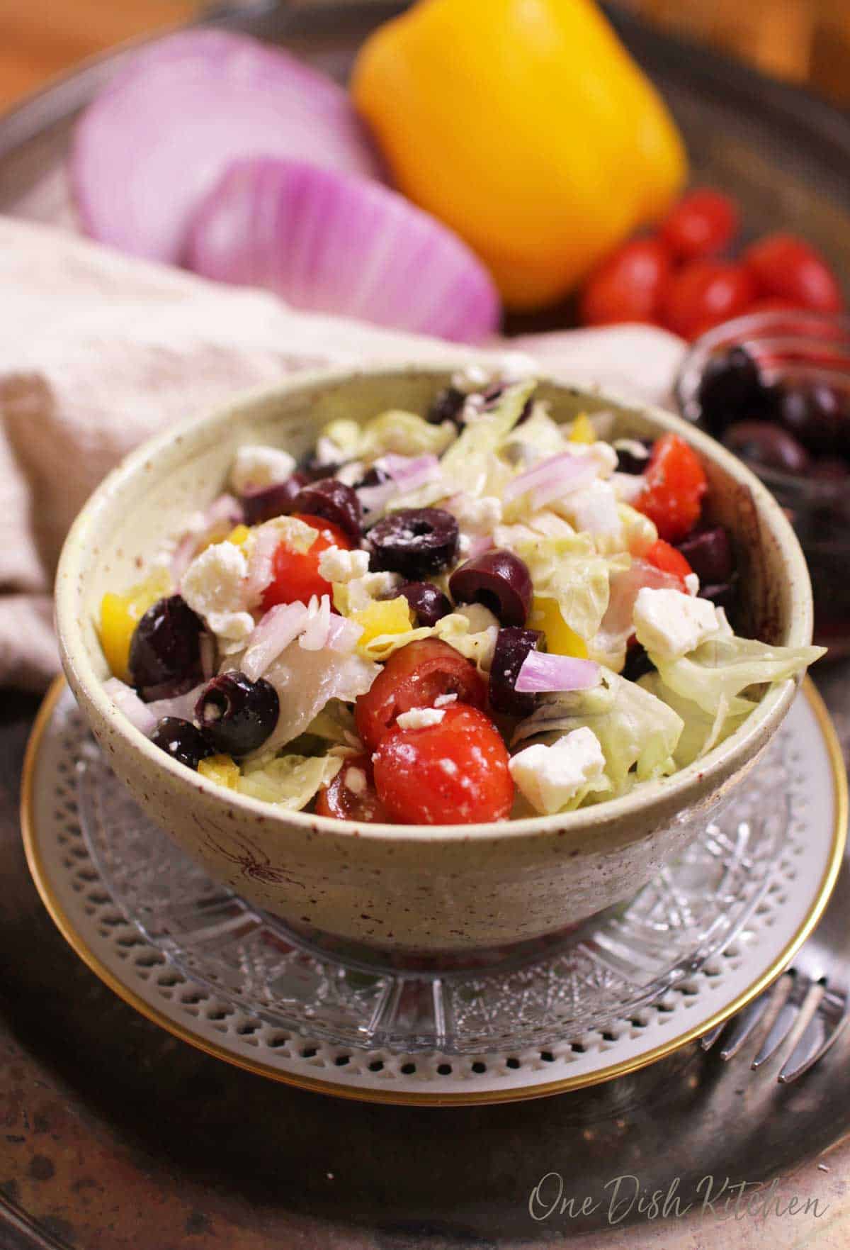 A Greek salad with olives, feta, tomatoes in a bowl with whole vegetables in the background.