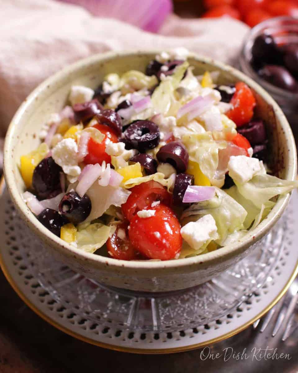 A Greek salad with olives, feta, tomatoes in a bowl with whole vegetables in the background.