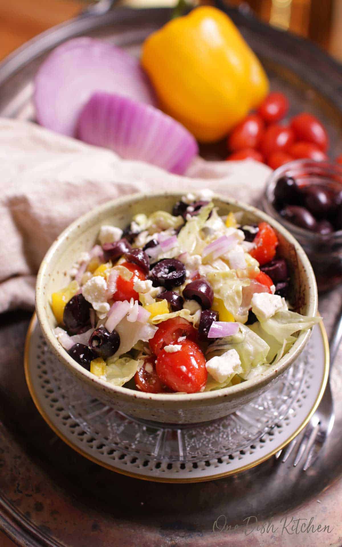 An overhead view of a Greek salad in a bowl on a silver tray next to a cloth napkin and an assortment of vegetables.