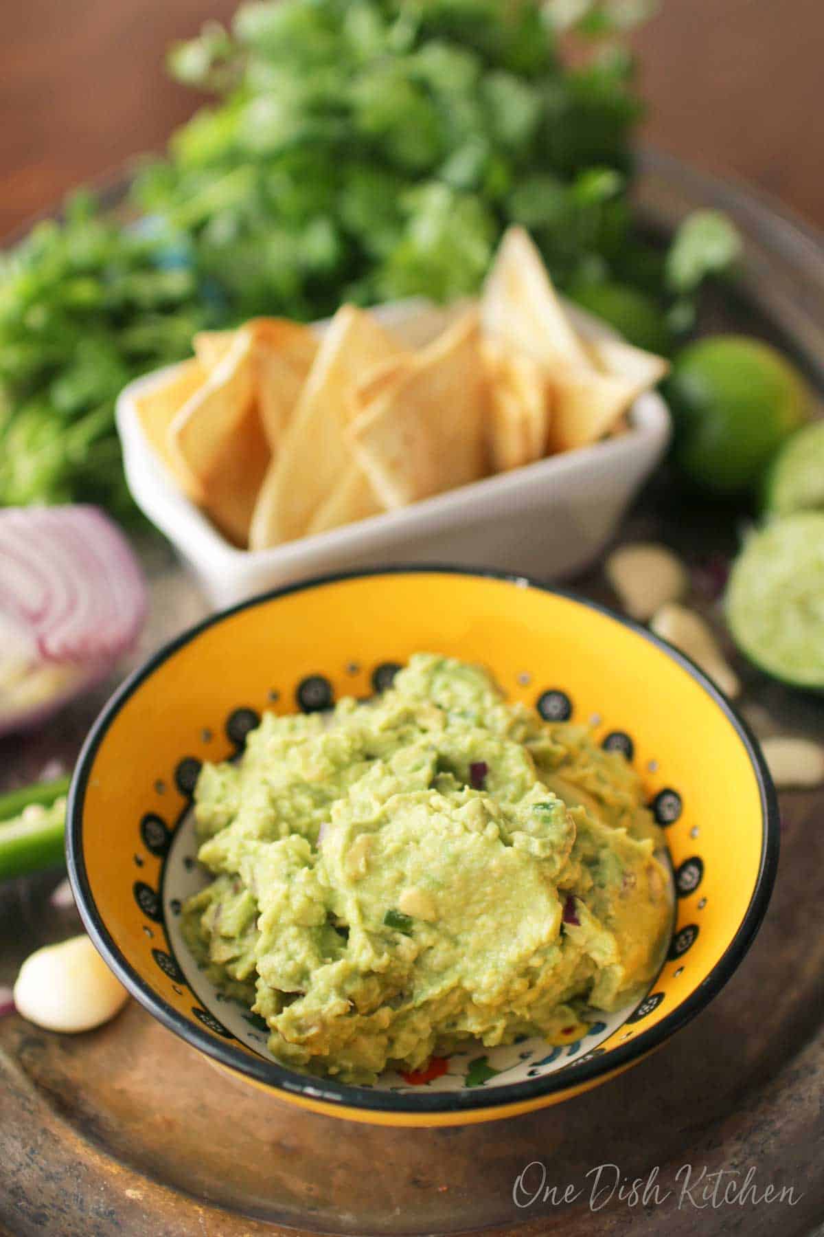 A small bowl of guacamole on a metal tray with a small bowl of homemade tortilla chips, an onion, garlic cloves, limes, and cilantro.
