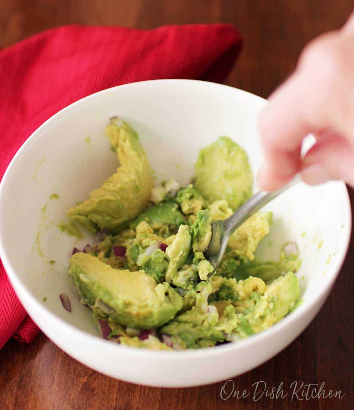 Mashing avocado, chopped red onions, garlic, and salt in a small bowl with a fork.
