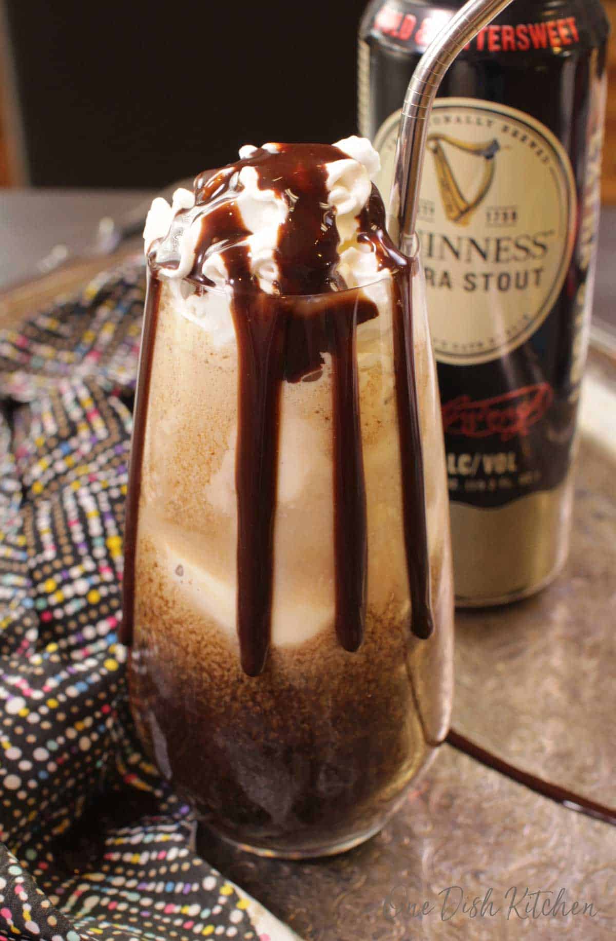 A Guinness Ice cream float made with vanilla ice cream in a glass with a metal straw, whipped cream, and chocolate syrup and a can of Guinness Stout  is in the background 