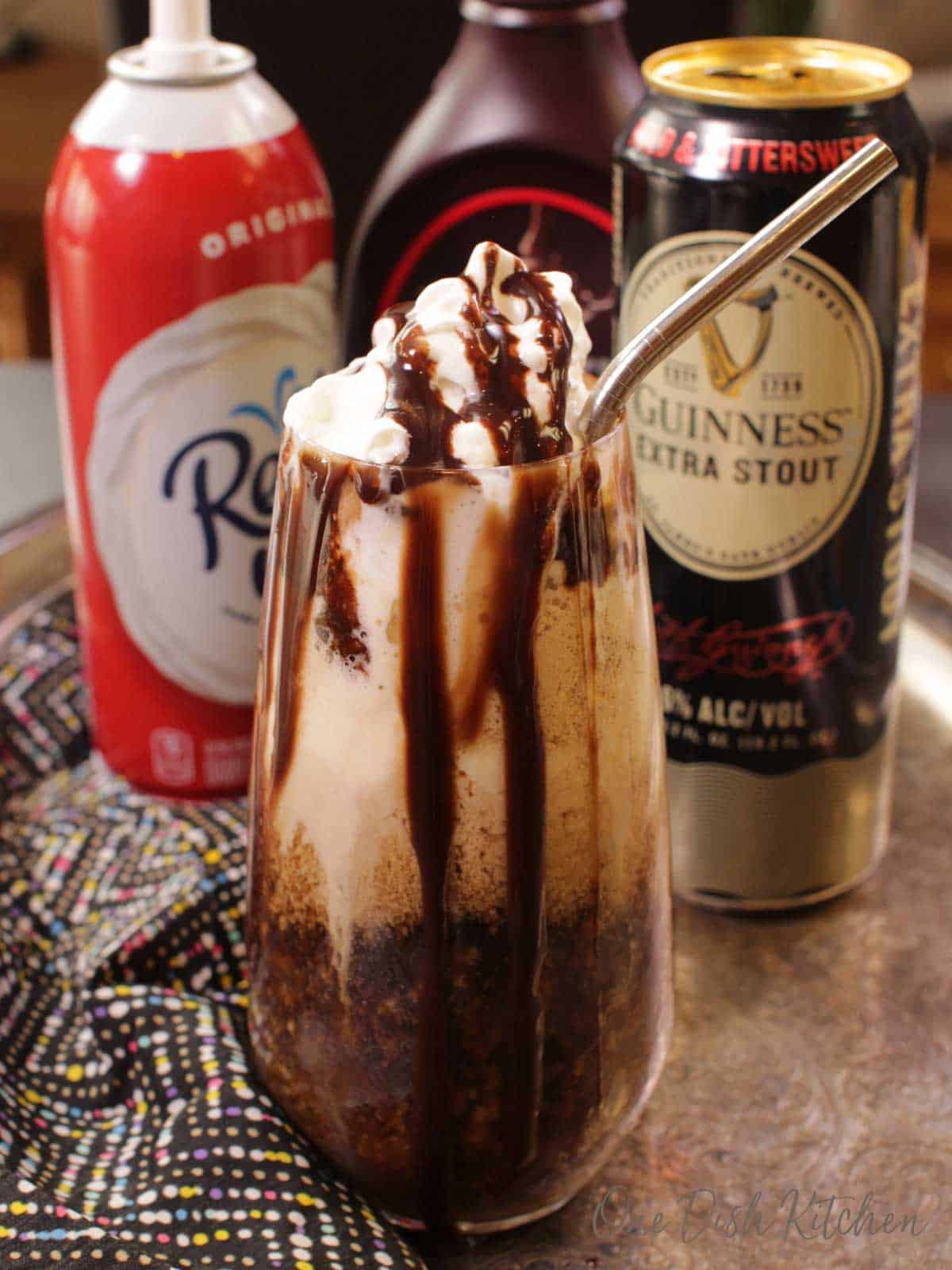 A Guinness Float made with vanilla ice cream and topped with a metal straw, whipped cream and chocolate syrup on a metal tray with a can of whipped cream, chocolate syrup, and a can of Guinness Stout