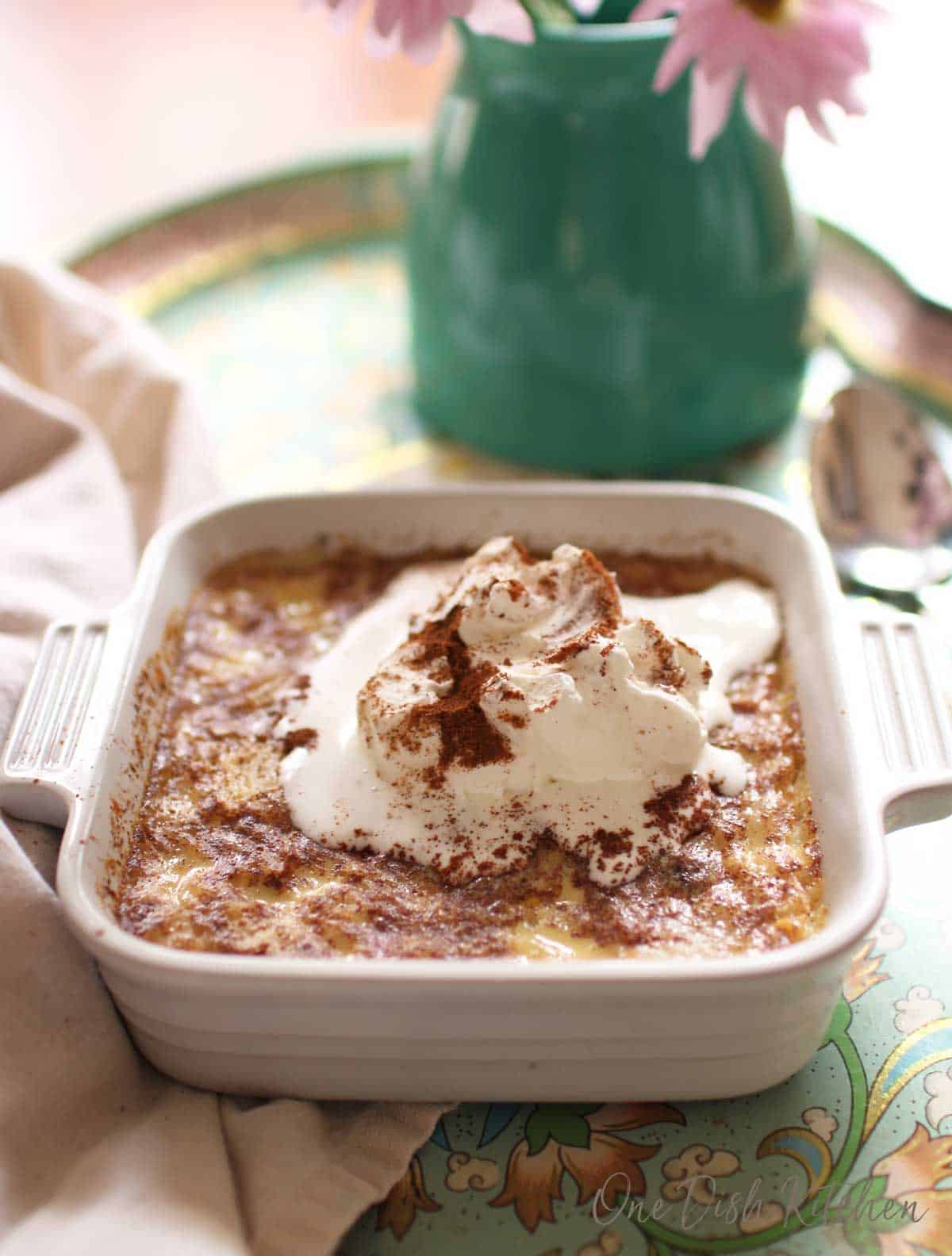 Baked rice pudding topped with whipped cream and ground cinnamon in a small baking dish on a tray 