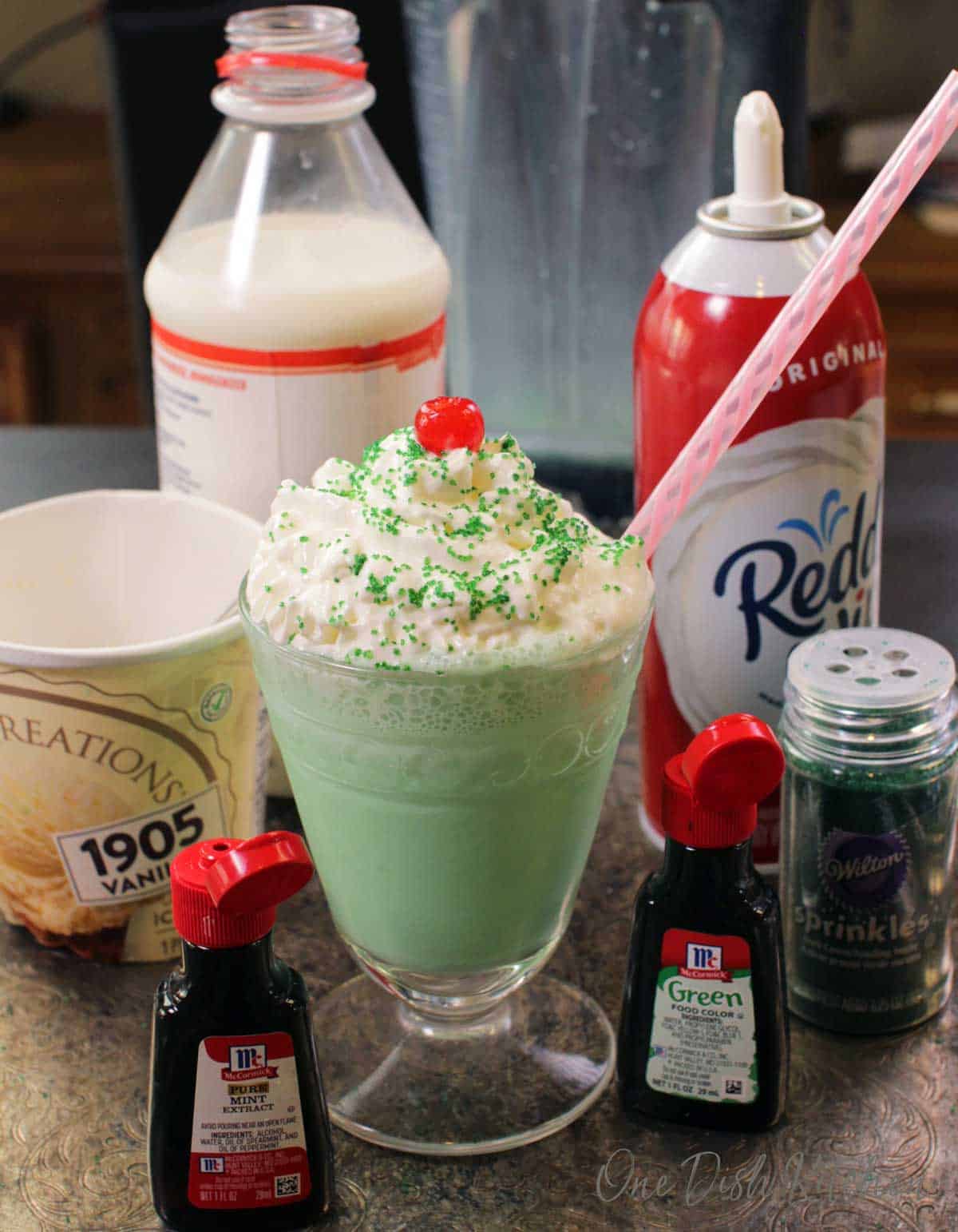 Ingredients in a shamrock shake on a tray- milk, whipped cream, vanilla ice cream, sprinkles, green food coloring, and a jar of mint extract.