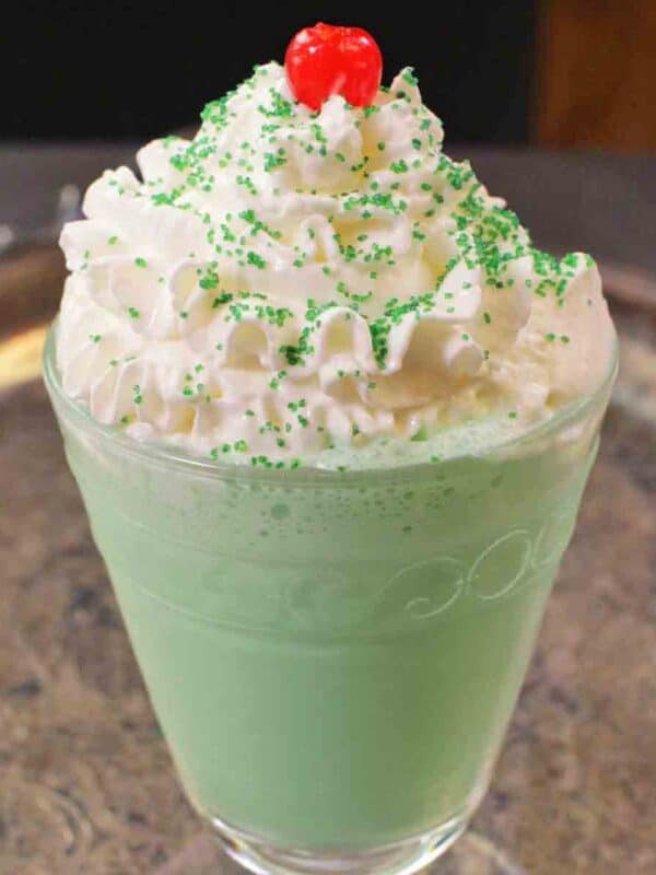 a green colored shamrock shake with whipped topping and cheery on top in a decorative glass.