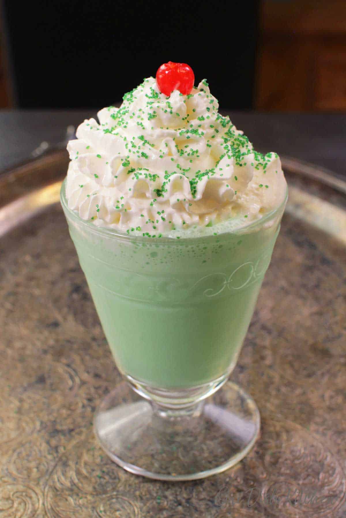A shamrock shake in a tall dessert glass topped with whipped cream, green sprinkles, and a maraschino cherry.