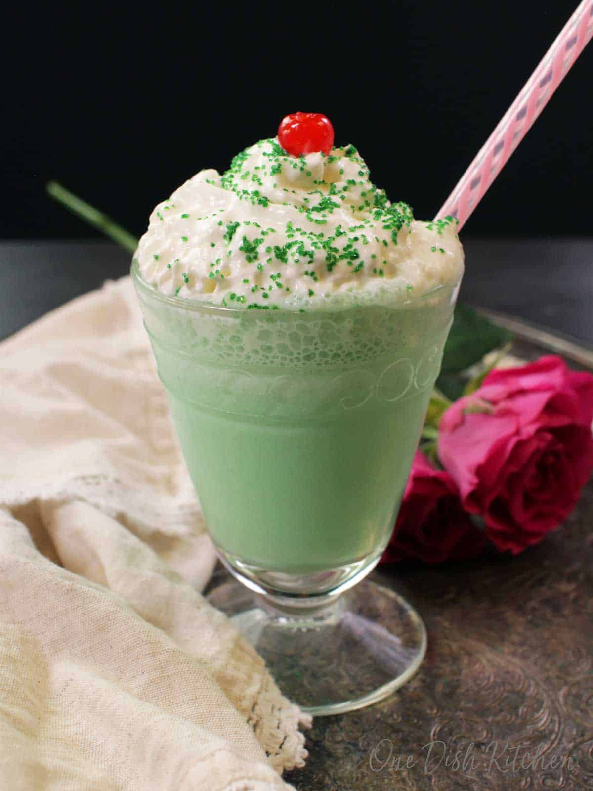 A shamrock shake in a tall dessert glass topped with whipped cream, green sprinkles, and a maraschino cherry.