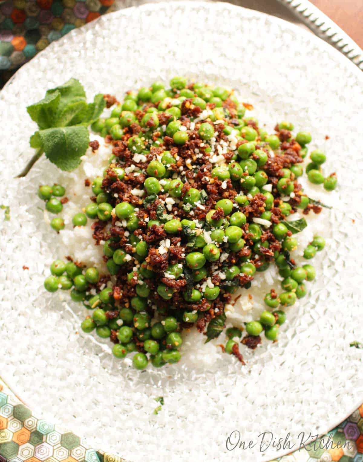 Overhead view of peas with chorizo, cotija cheese crumbles, and mint leaves over white rice on a clear glass plate.