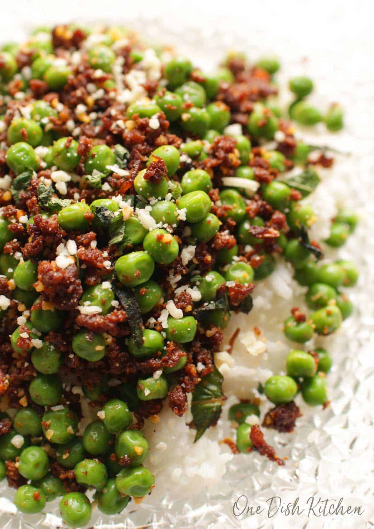 Closeup of peas with chorizo, cotija cheese crumbles, and mint leaves over white rice on a clear glass plate 