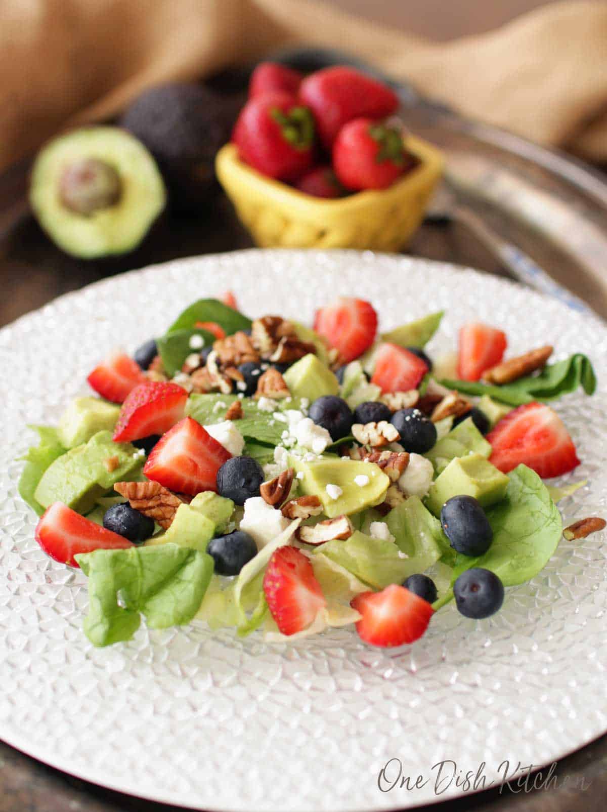 A salad of avocados, strawberries, blueberries, pecans, feta cheese, and spinach plated on a metal tray with a small bowl of strawberries and half of an avocado 