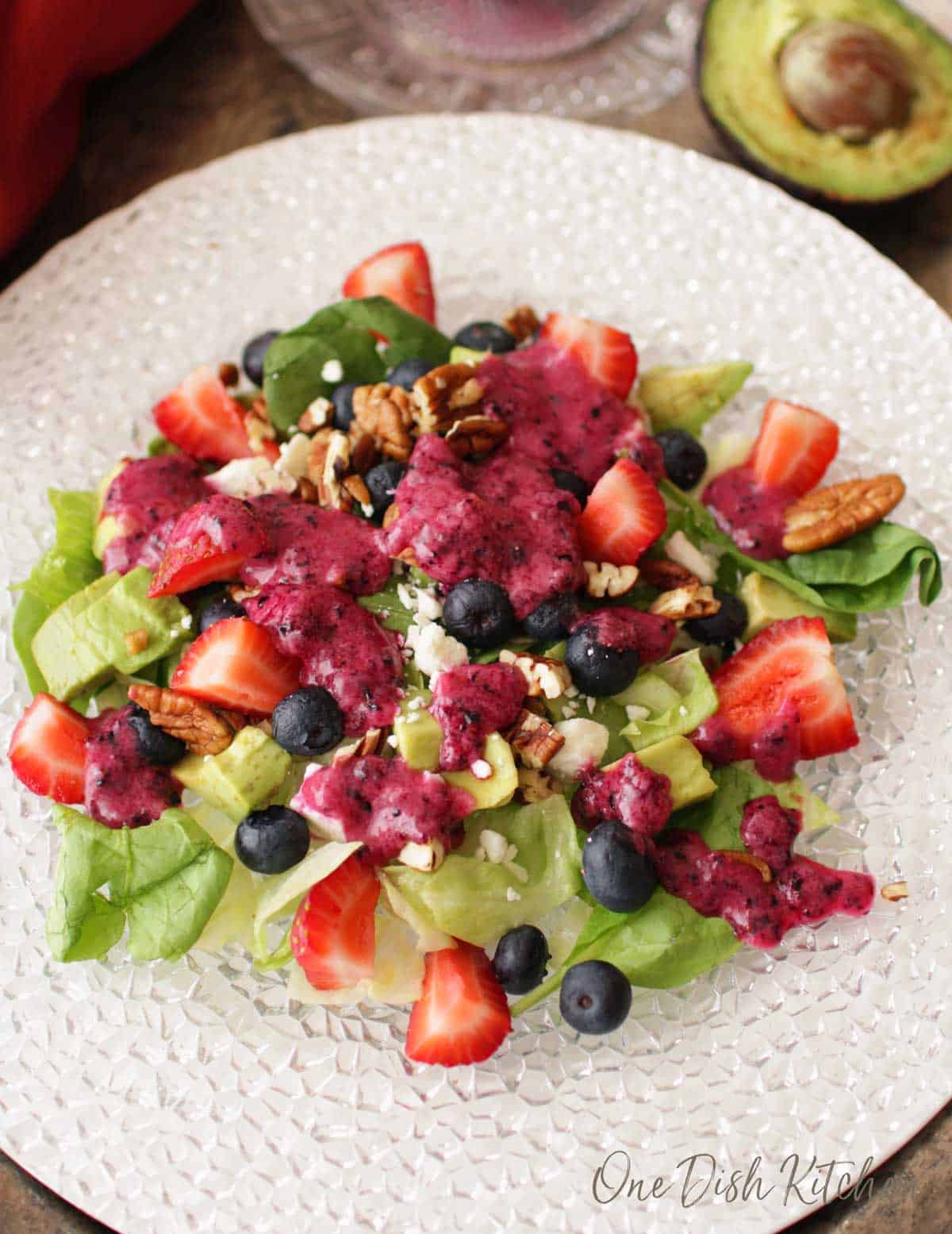 An overhead view of a salad consisting of avocados, strawberries, blueberries, pecans, feta cheese, and spinach with blueberry vinaigrette drizzled over the top
