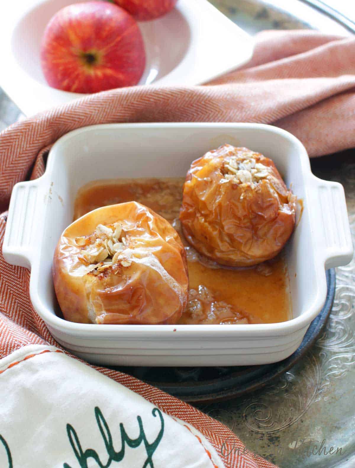 Two baked apples in a small baking dish filled with oats and brown sugar next to a bowl of apples