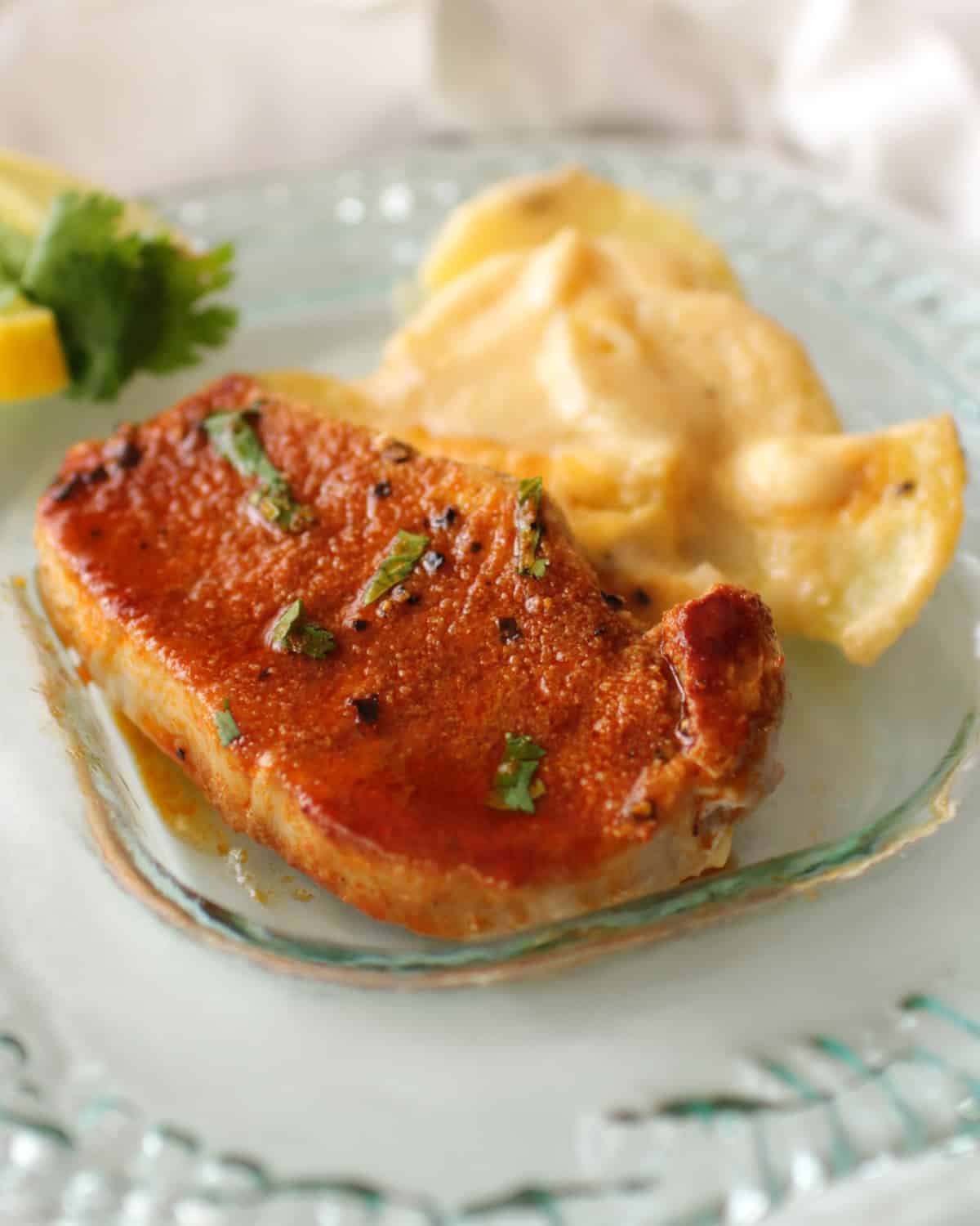 one baked pork chop on a clear plate next to scalloped potatoes and lemon slices.