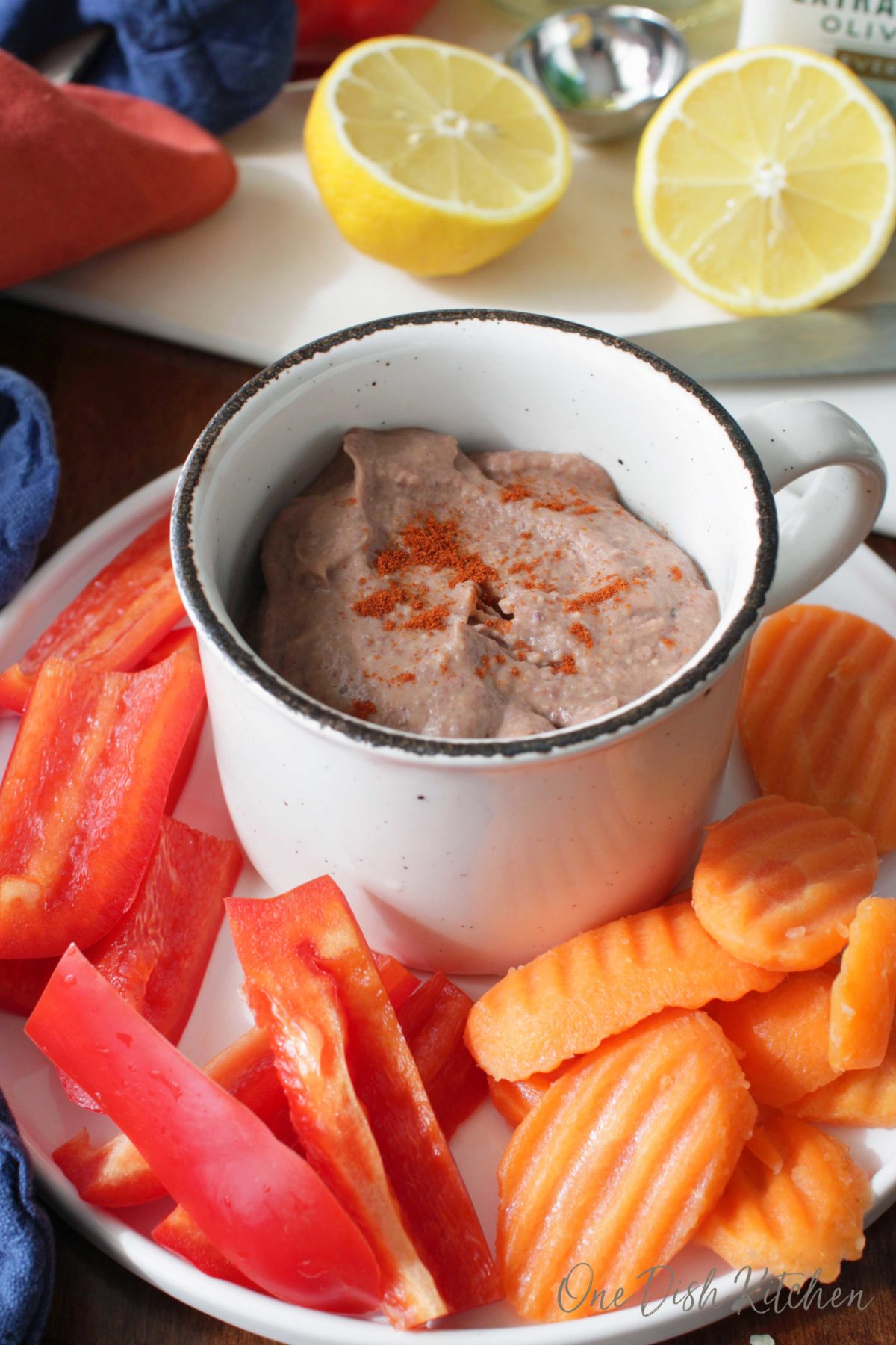 A bowl of black bean hummus plated with sliced carrots and red peppers with lemon halves in the background.