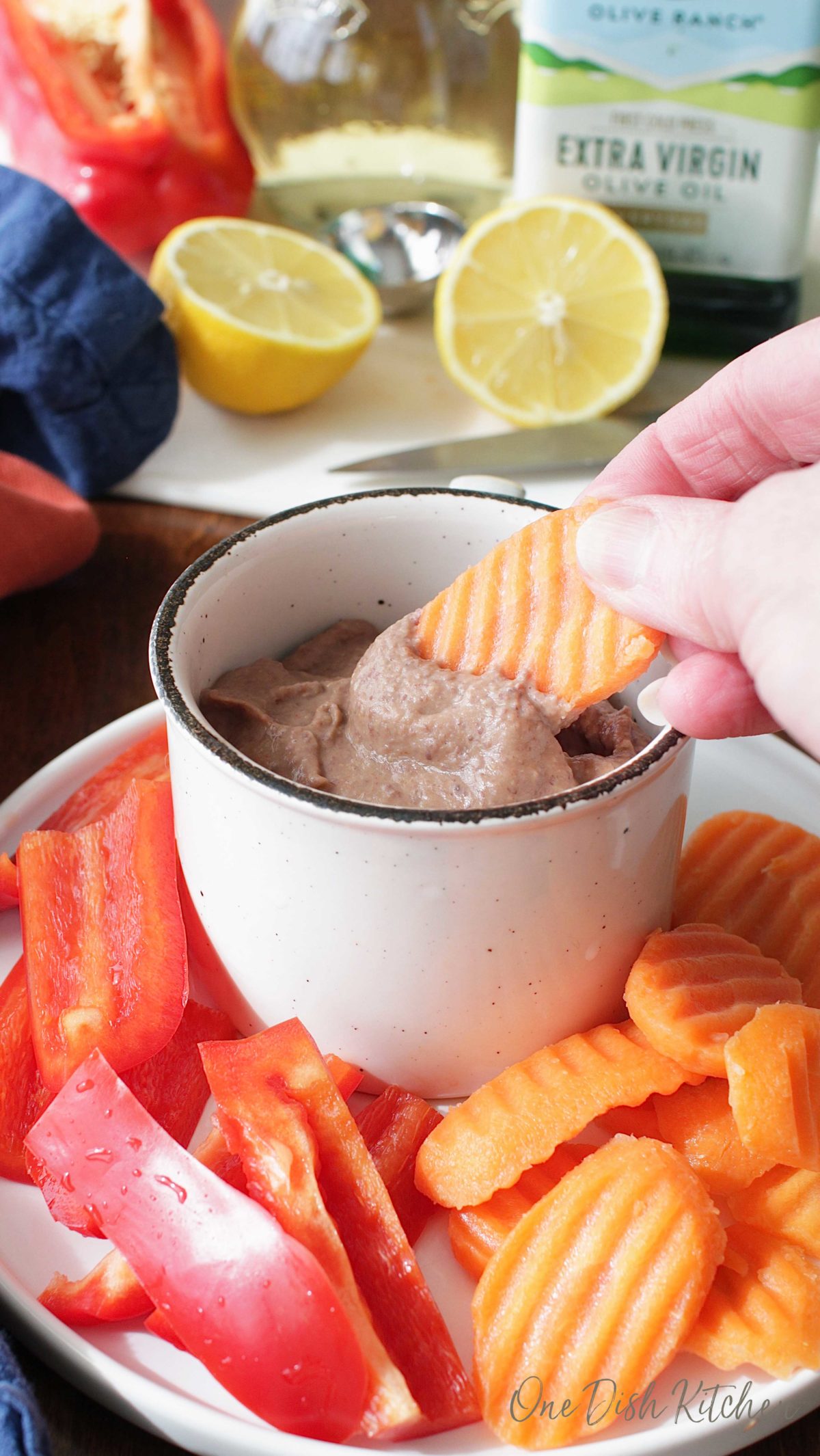 Carrot dipped in a small bowl of black bean hummus plated with sliced carrots and red peppers.