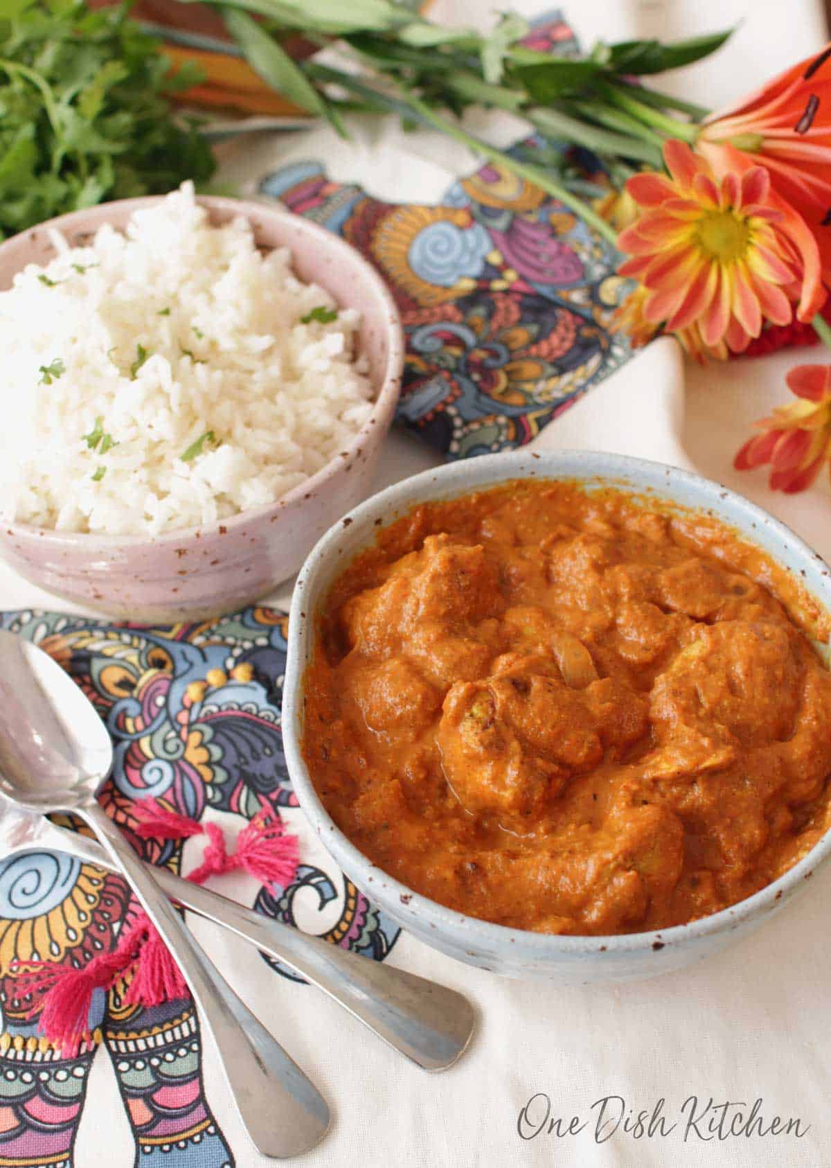 a bowl of butter chicken next to a bowl of white rice on a flowered tablecloth
