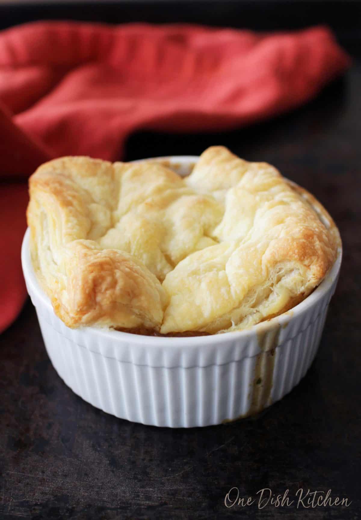 A single serving chicken pot pie topped with a puff pastry crust in a small ramekin next to a red cloth napkin. 