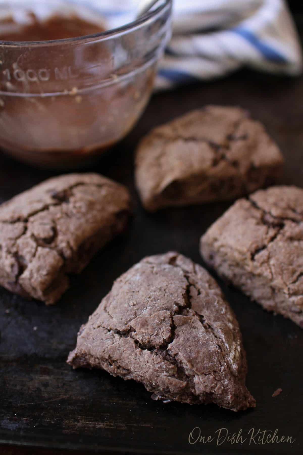 Chocolate scones on a baking sheet with a bowl of melted chocolate next to the scones.