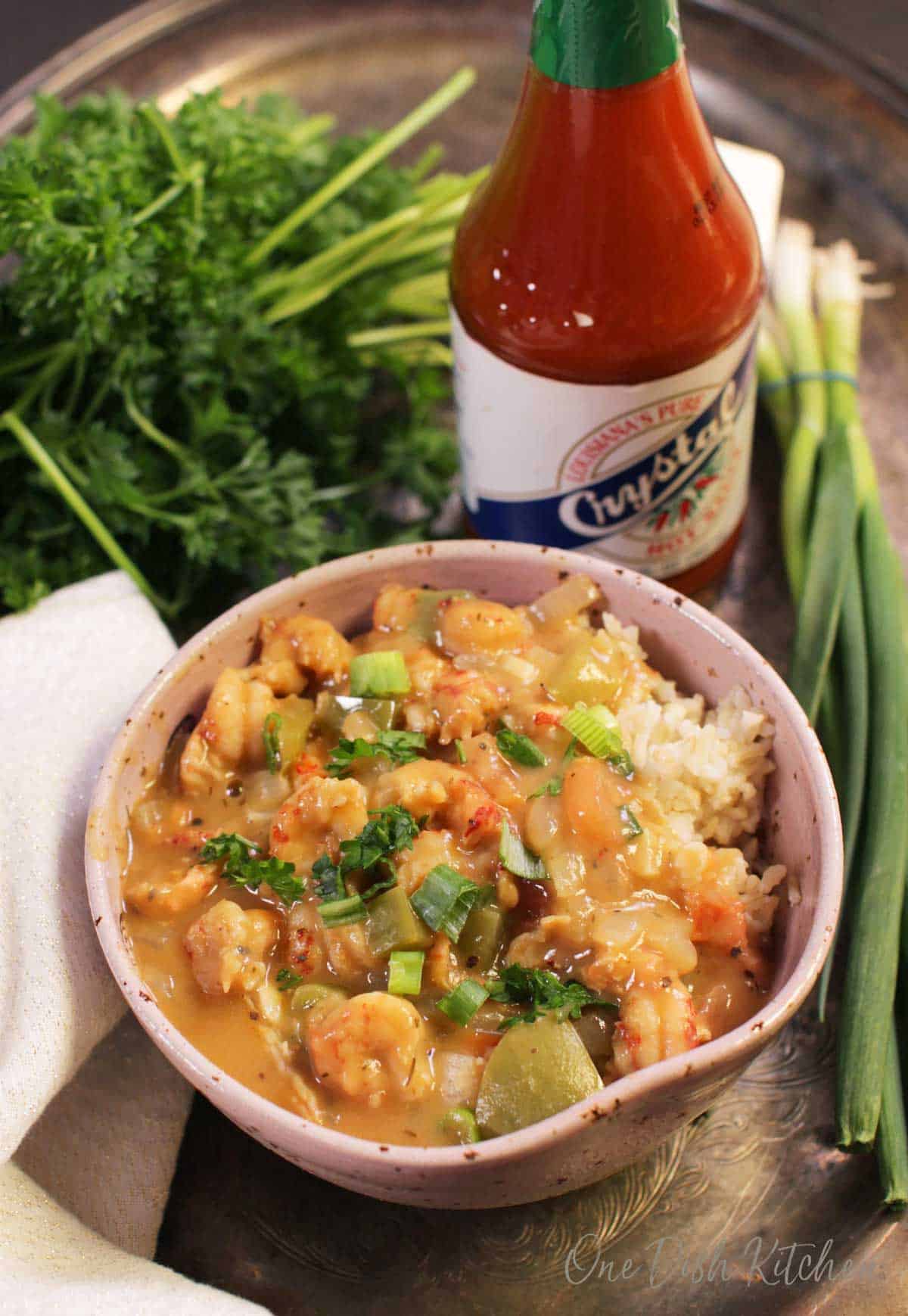 An overhead view of a bowl of crawfish Étouffée with white rice on a metal tray next to a bottle of Louisiana hot sauce and green onions