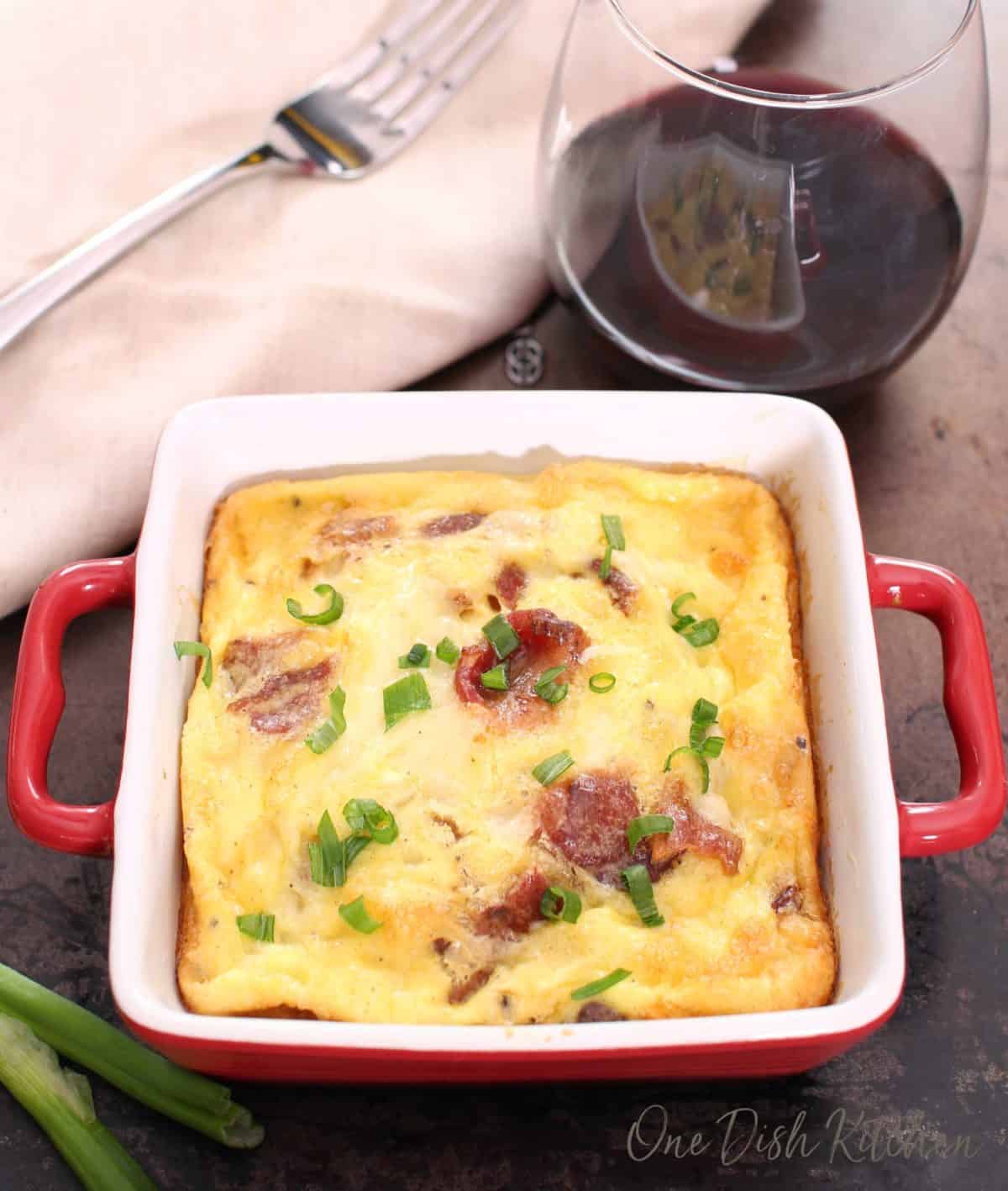 Overhead view of a crustless quiche with bacon and cheese in a small baking dish on a tray with a glass of red wine and a fork.