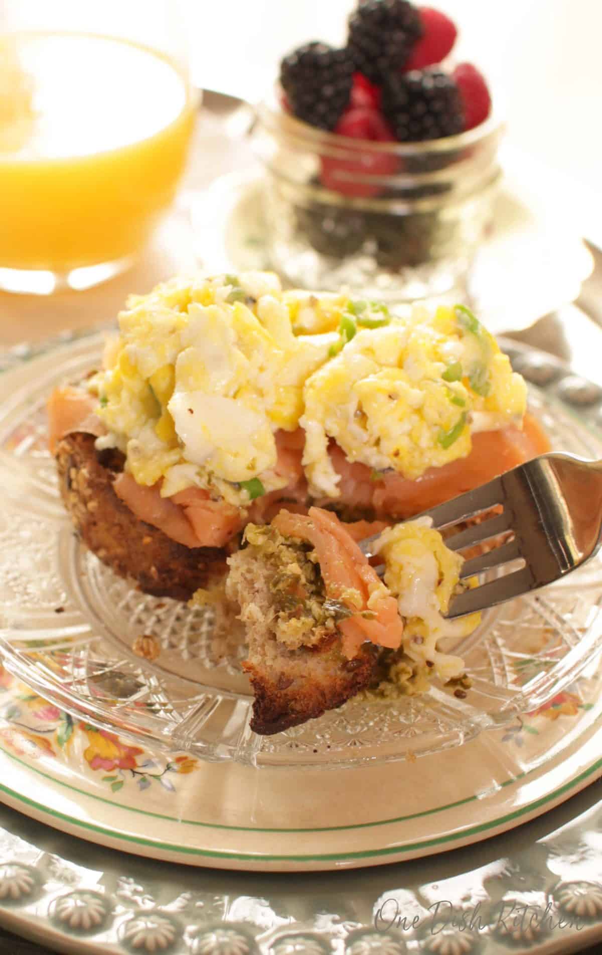 A closeup of a forkful of french bread topped with smoked salmon, pesto, and scrambled eggs next to a glass of orange juice and a jar of blackberries and raspberries