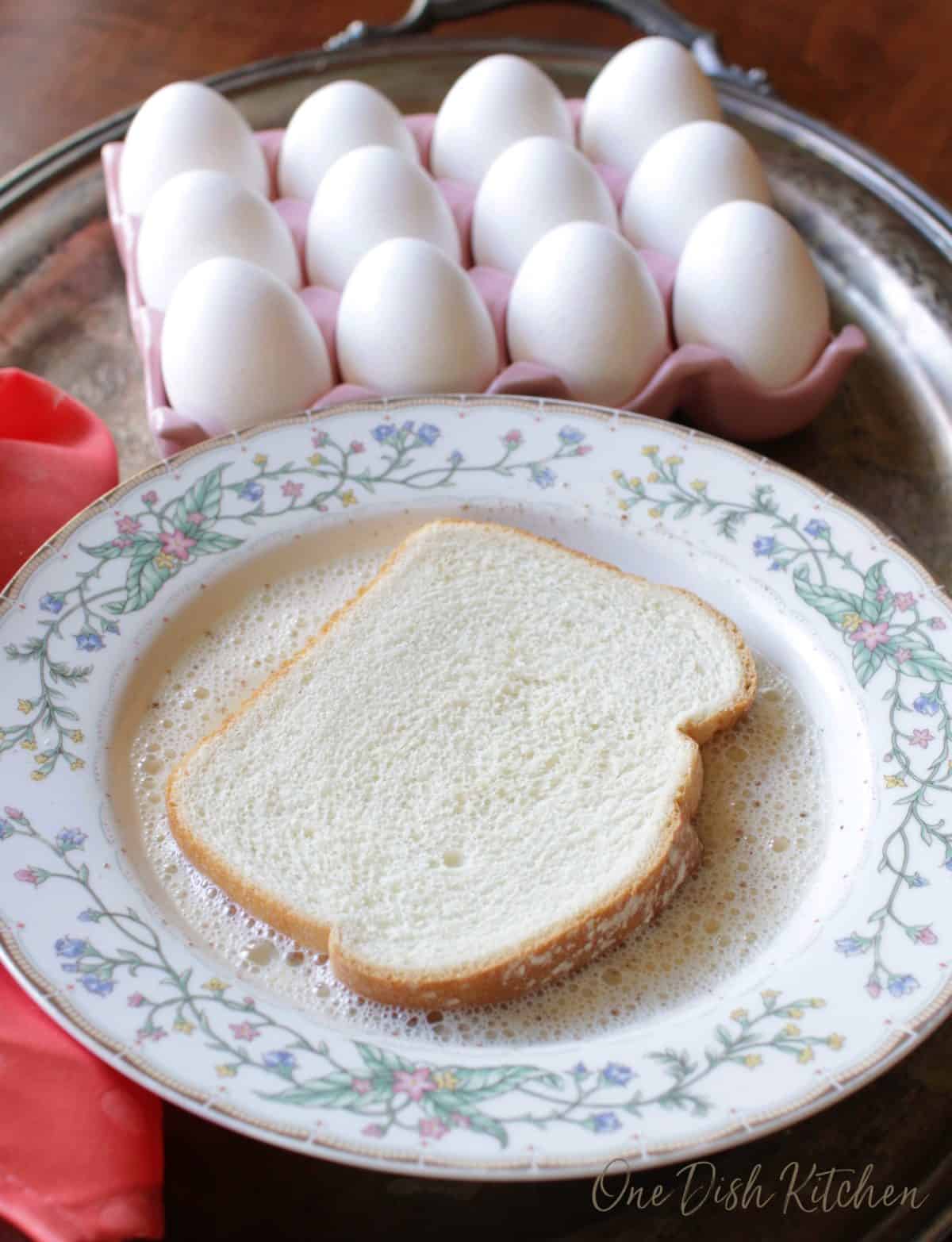 A slice of white bread soaking in milk before making french toast on a metal tray with a tray of a dozen eggs and a red cloth napkin