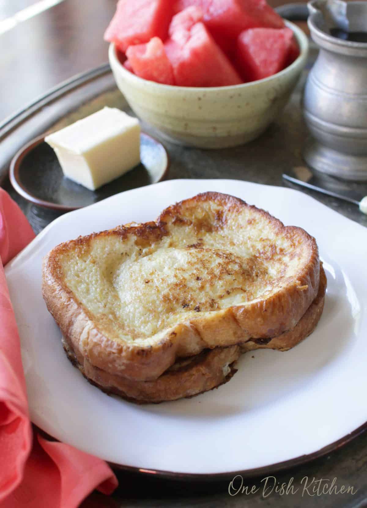 A stack of two french toast slices on a plate next to a stick of butter and a bowl of watermelon slices all on a metal tray