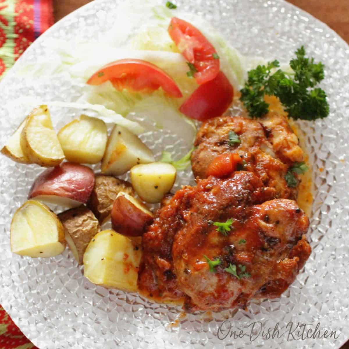 An overhead view of italian chicken with a small salad of iceberg lettuce and tomato slices and chopped roasted potatoes on a plate.