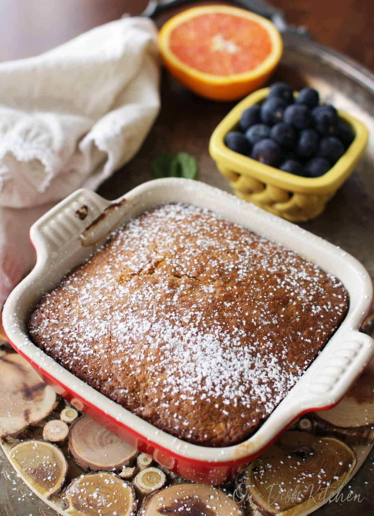 A mini oatmeal cake in a small baking dish topped with powdered sugar on a wooden trivet next to half an orange and a small bowl of blueberries all on a metal tray.