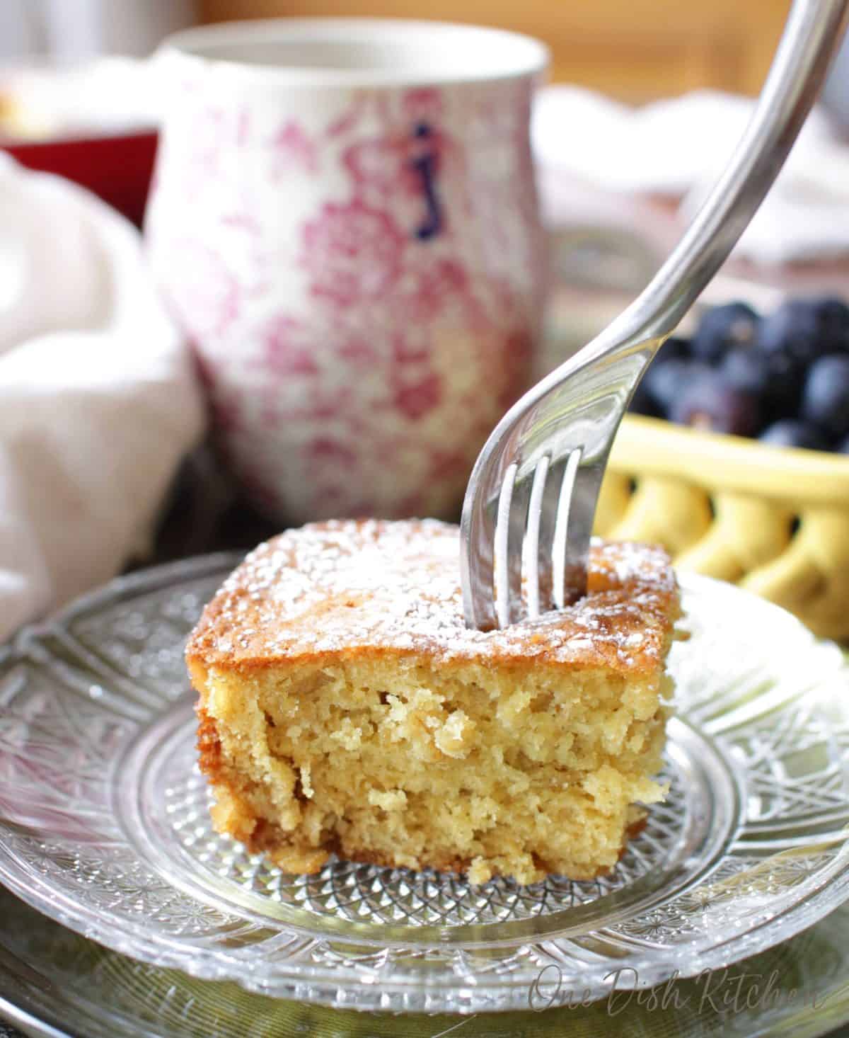 A fork piercing the top of a slice of orange oatmeal cake topped with powdered sugar next to a coffee mug and a small bowl of blueberries.