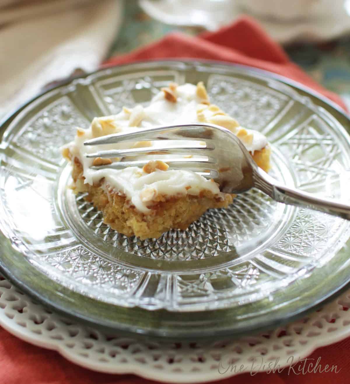 One slice of pineapple sheet cake topped with vanilla icing and chopped peanuts being eaten with a fork on a clear plate.