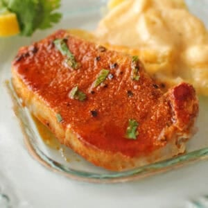 one baked pork chop on a clear plate next to scalloped potatoes and lemon slices.
