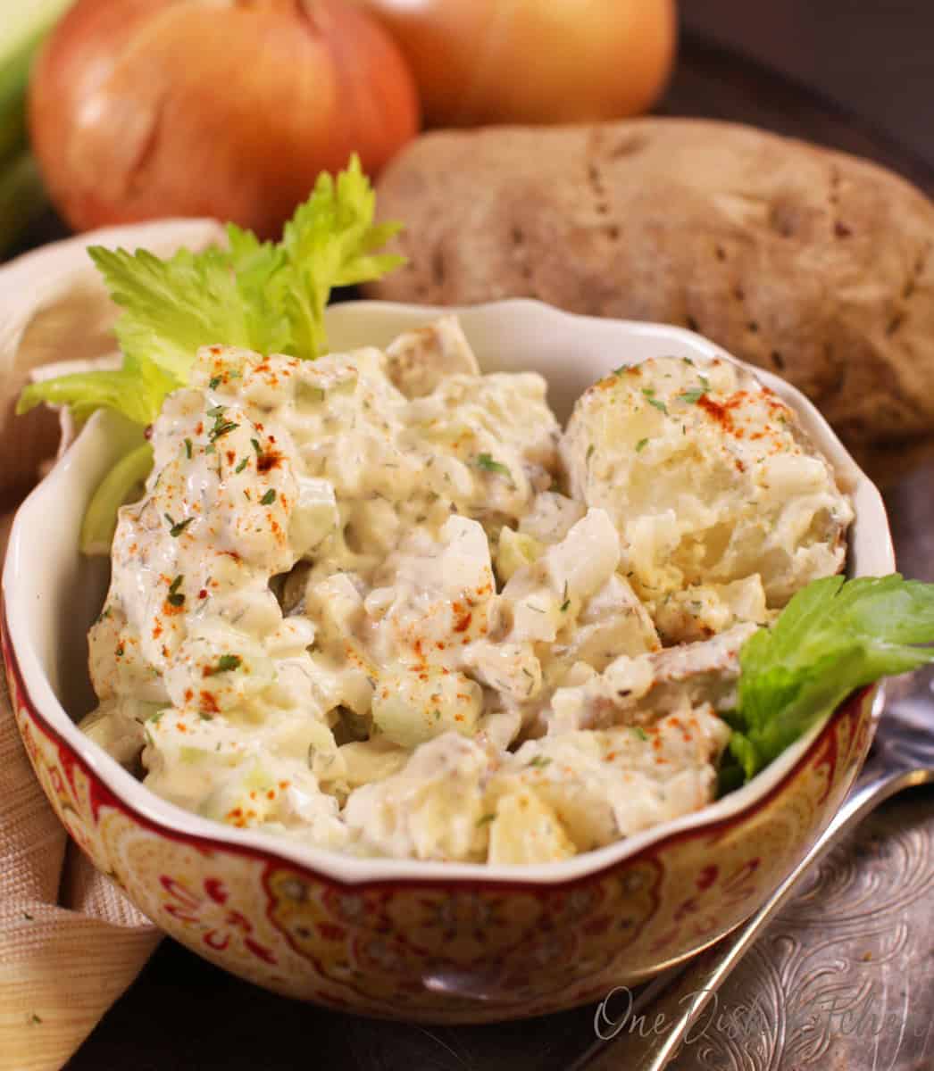 A bowl of potato salad next to a fork, a russet potato, and two onions all on a metal tray.