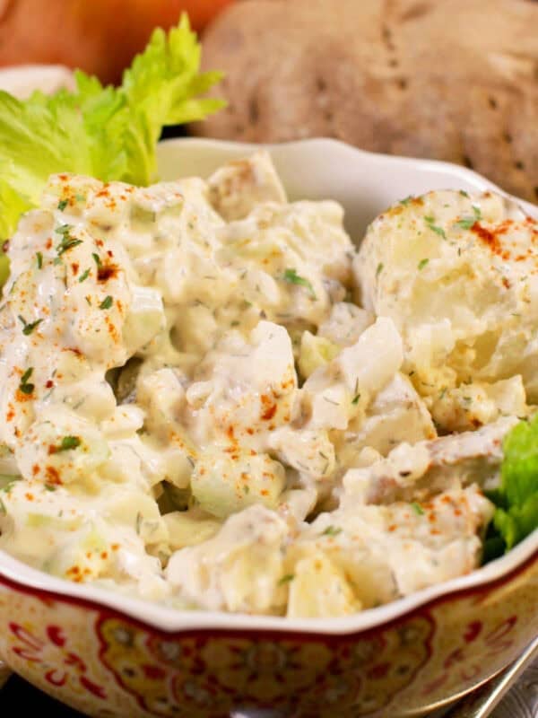 a red bowl filled with potato salad next to a russet potato and an onion.