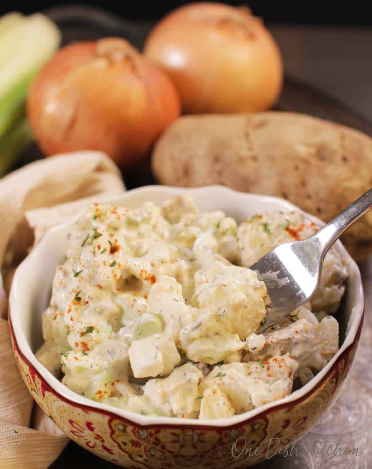A fork filled with potato salad from a small bowl next to a russet potato, and two onions all on a metal tray