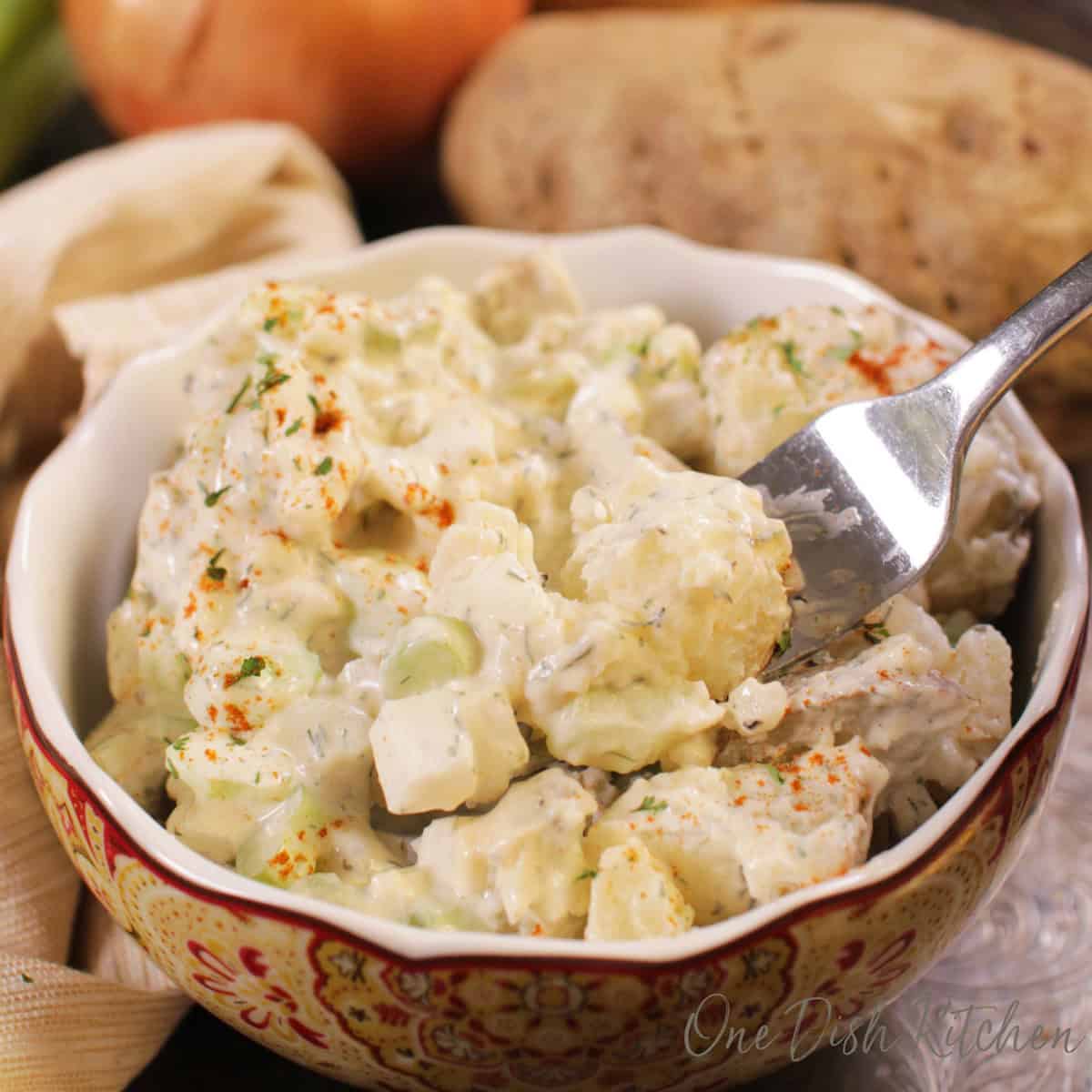 A fork filled with potato salad from a small bowl next to a russet potato, and two onions all on a metal tray.