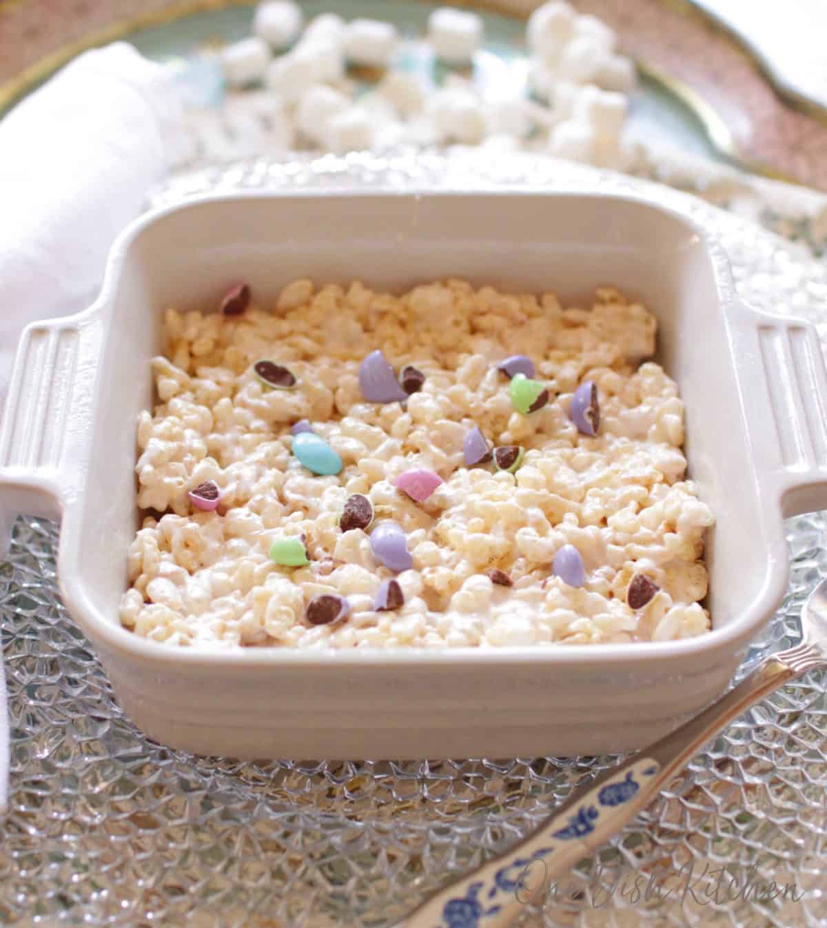 A giant rice krispies treat topped with pastel colored chocolate candies in a small baking dish next to a fork.