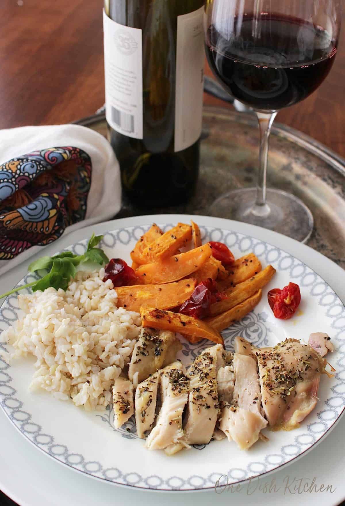 A plate of sliced chicken with roasted sweet potatoes slices, roasted cherry tomatoes, and brown rice next to a glass of red wine all on a metal tray