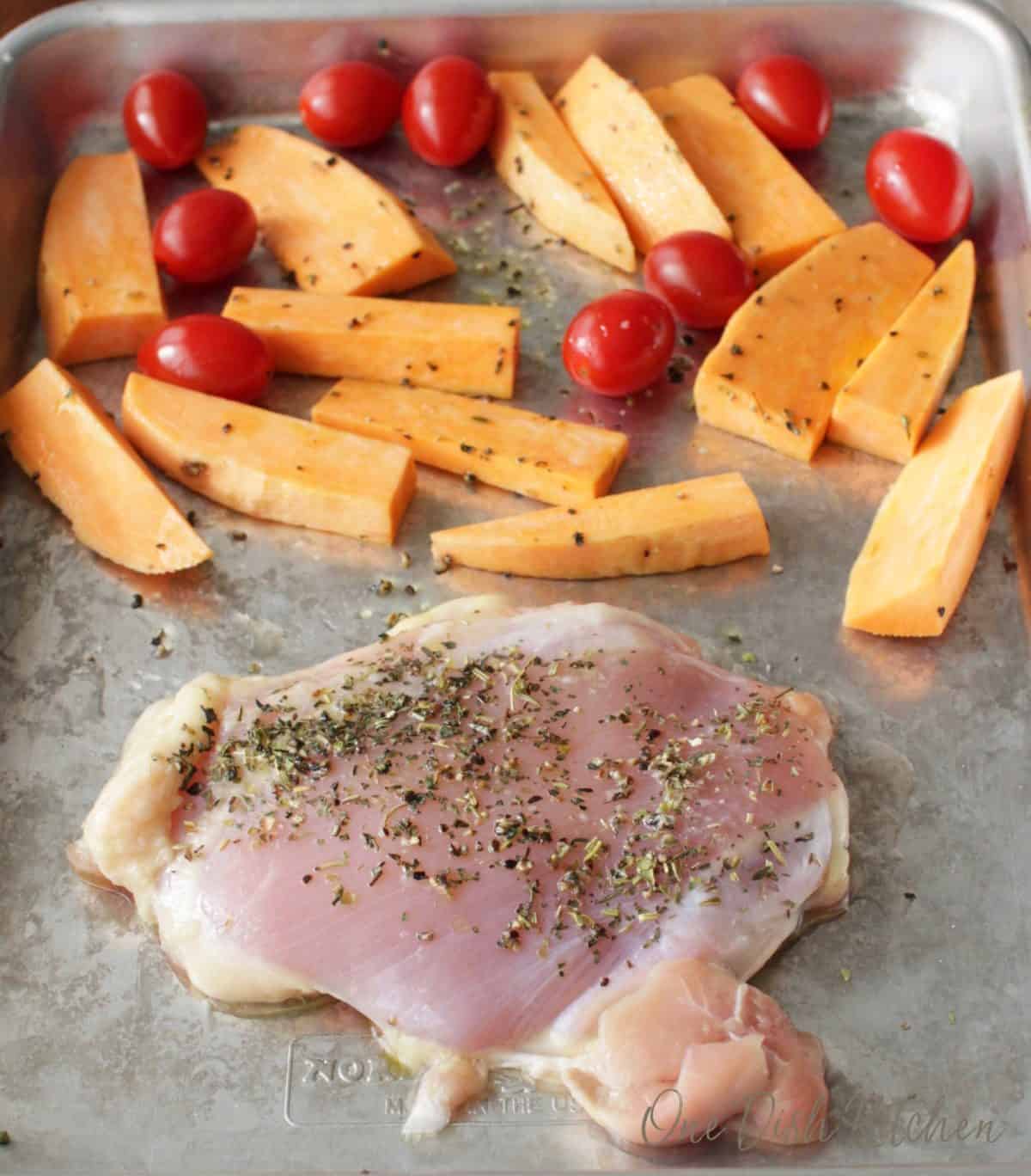 Raw chicken seasoned on a sheet pan along with sweet potato slices and cherry tomatoes 