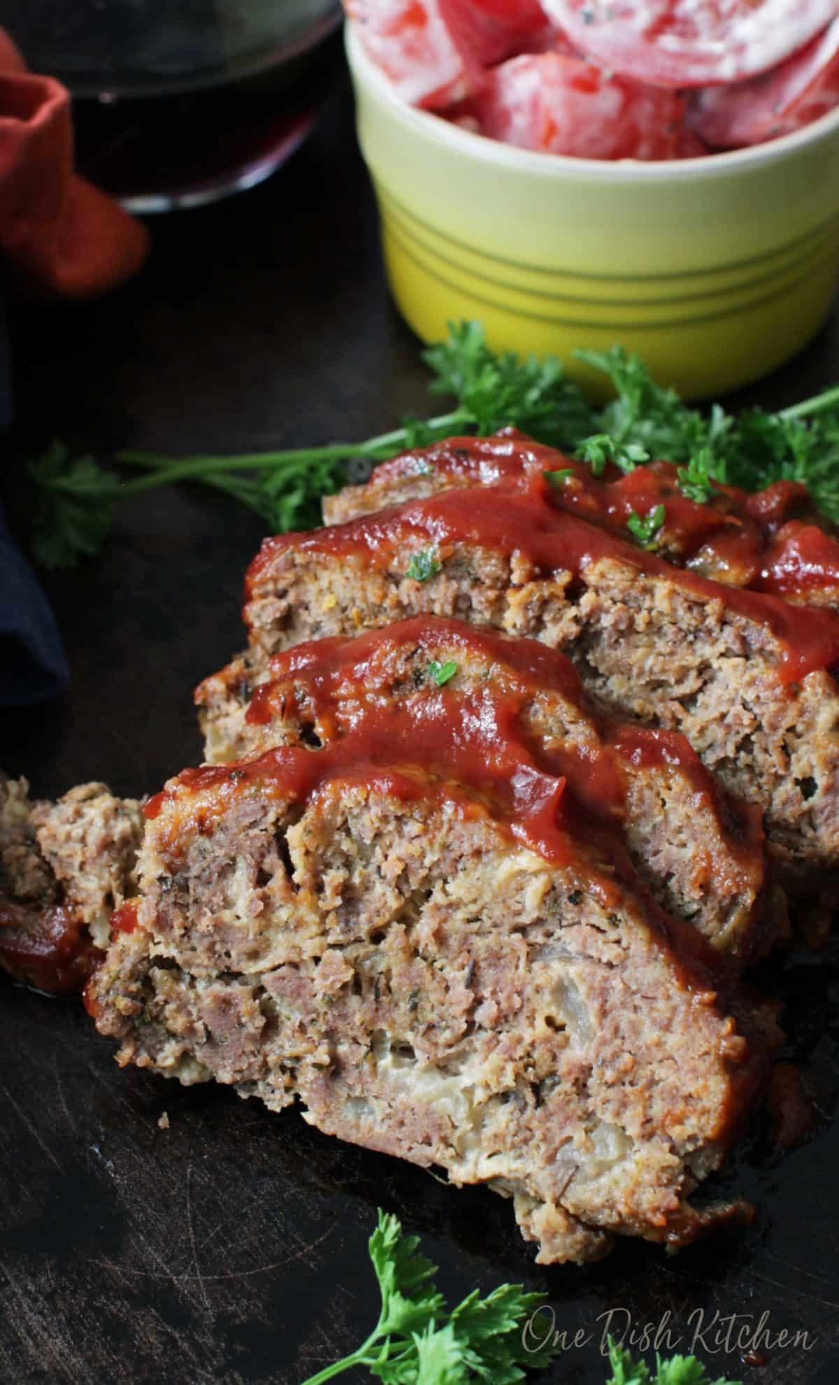 Meatloaf sliced into four slices on a dark wooden tray next to a small bowl of tomato slices with ranch dressing.