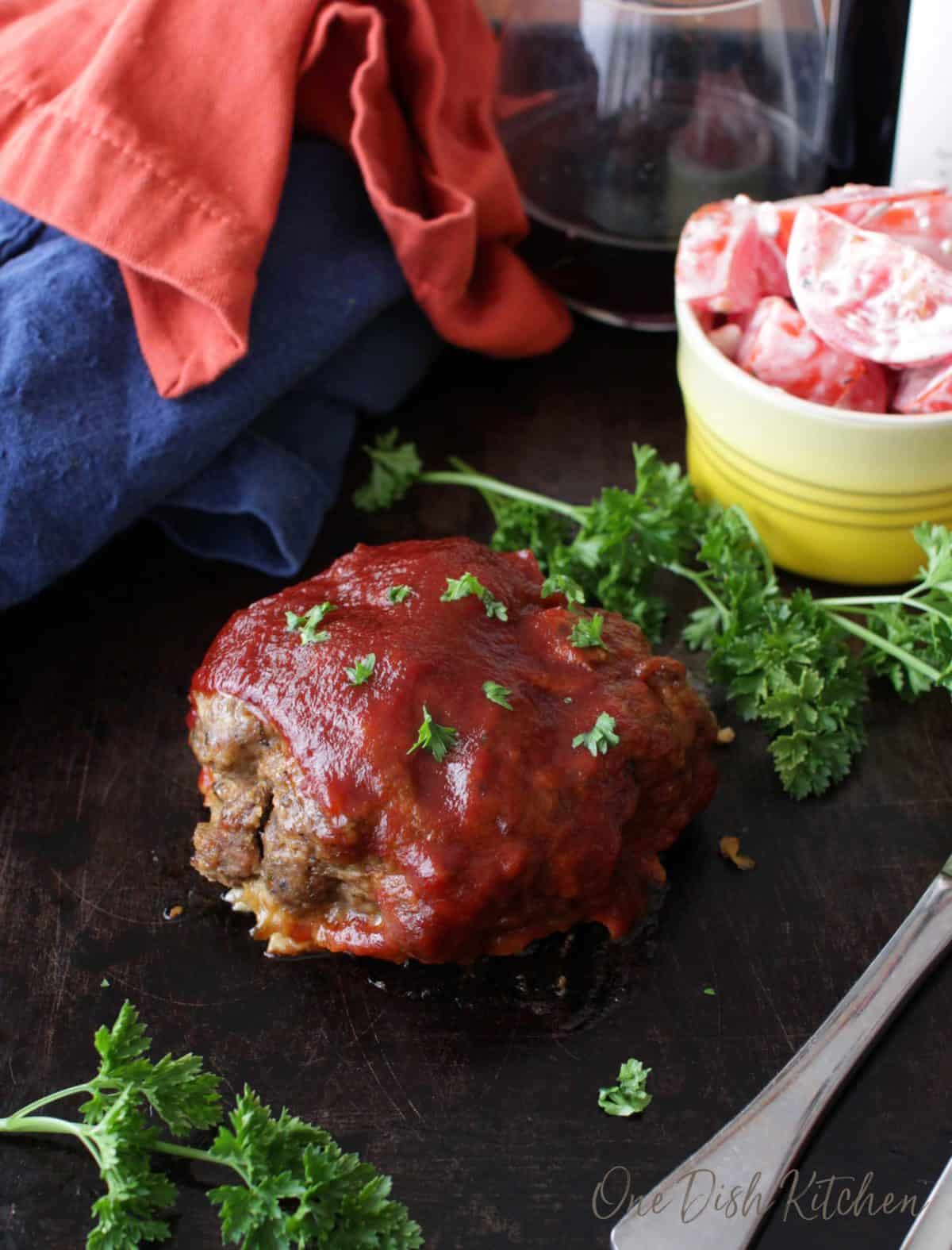A single serving of meatloaf on a dark wooden tray with a glass of red wine and a small bowl of tomato slices with ranch dressing.