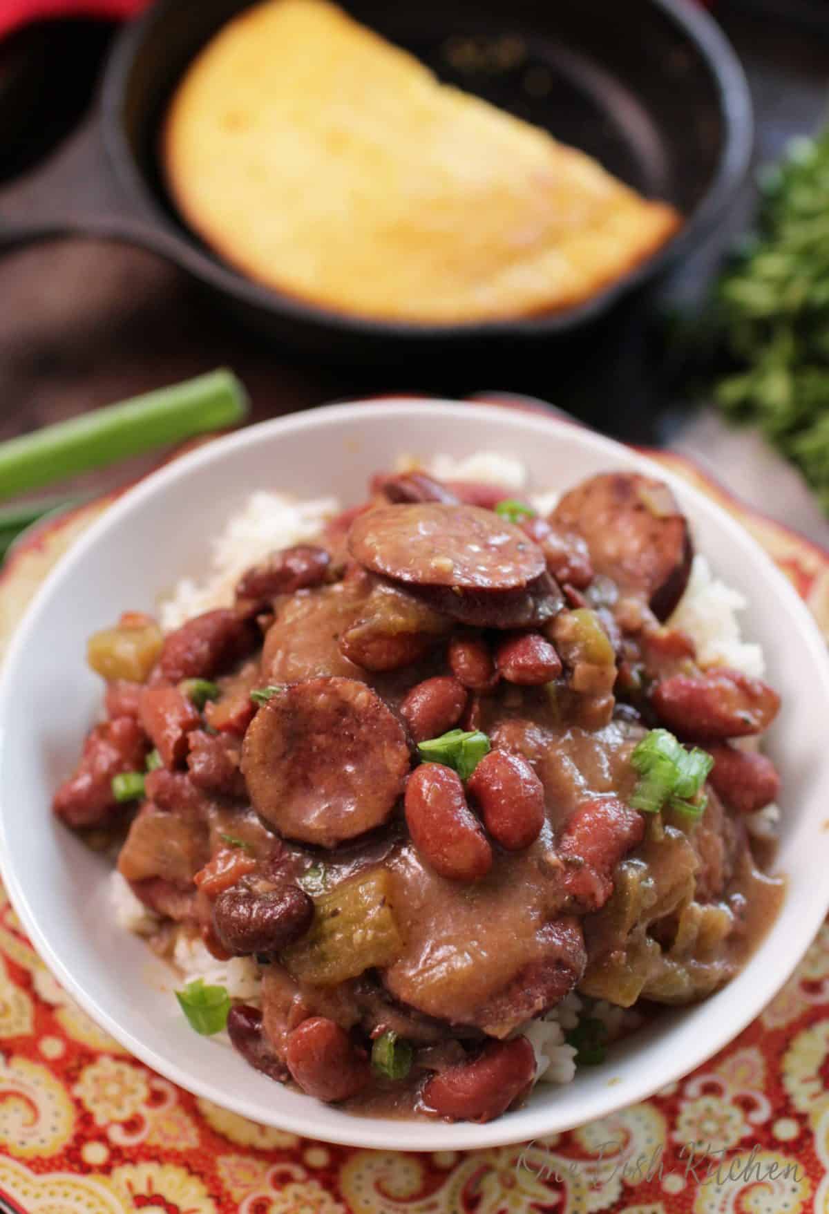 An overhead view of a bowl of red beans and white rice with sausage and cornbread in a cast iron skillet in the background.