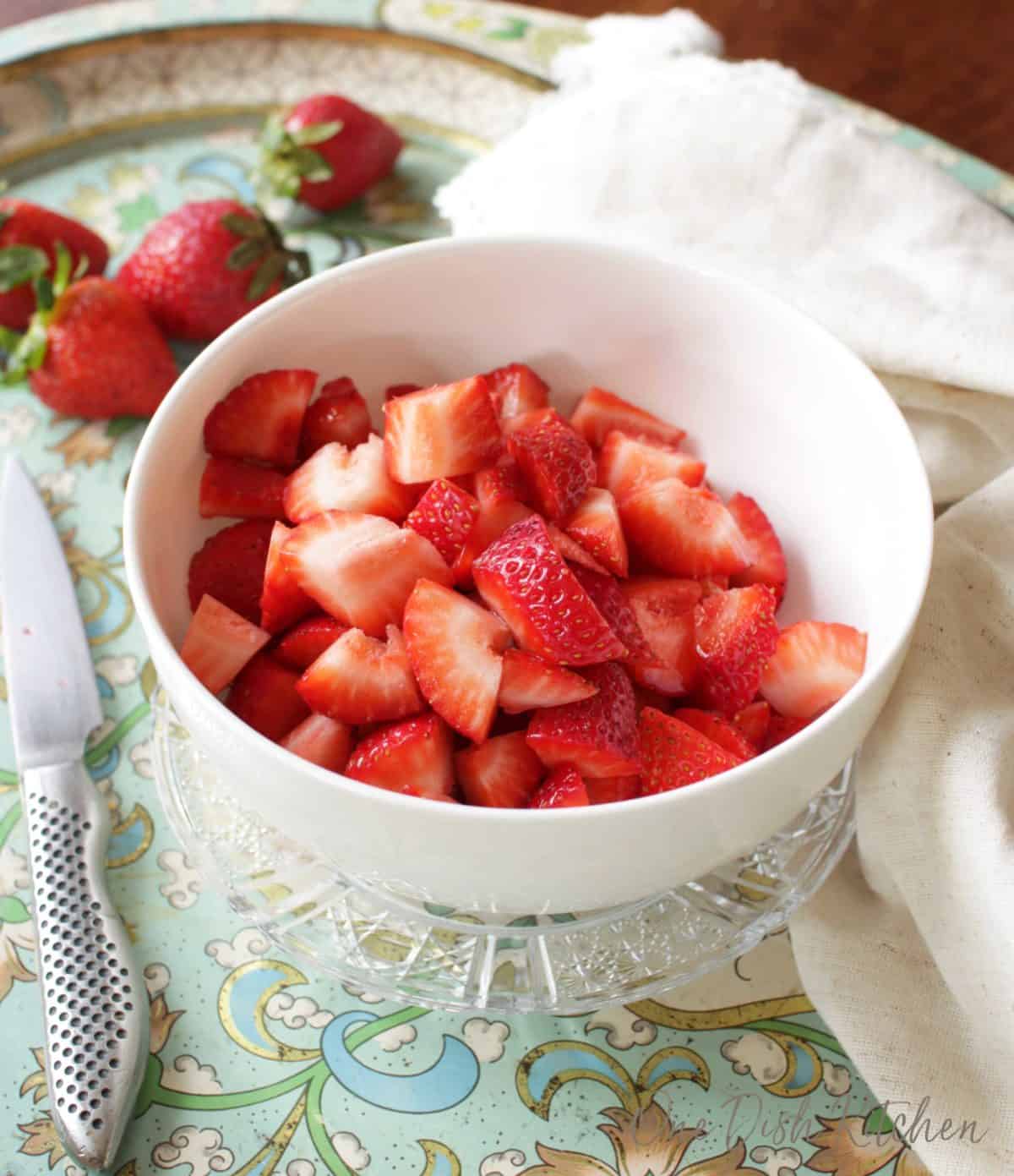 A bowl of fresh sliced strawberries plated next to a knife and scattered strawberries.