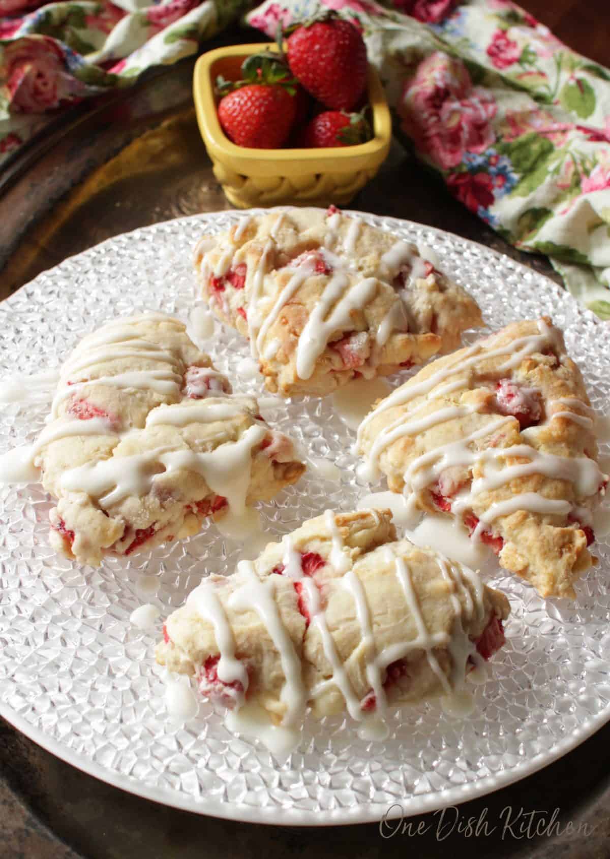 A plate of four strawberry scones with a drizzle of melted white chocolate on a plate next to a small bowl of strawberries 