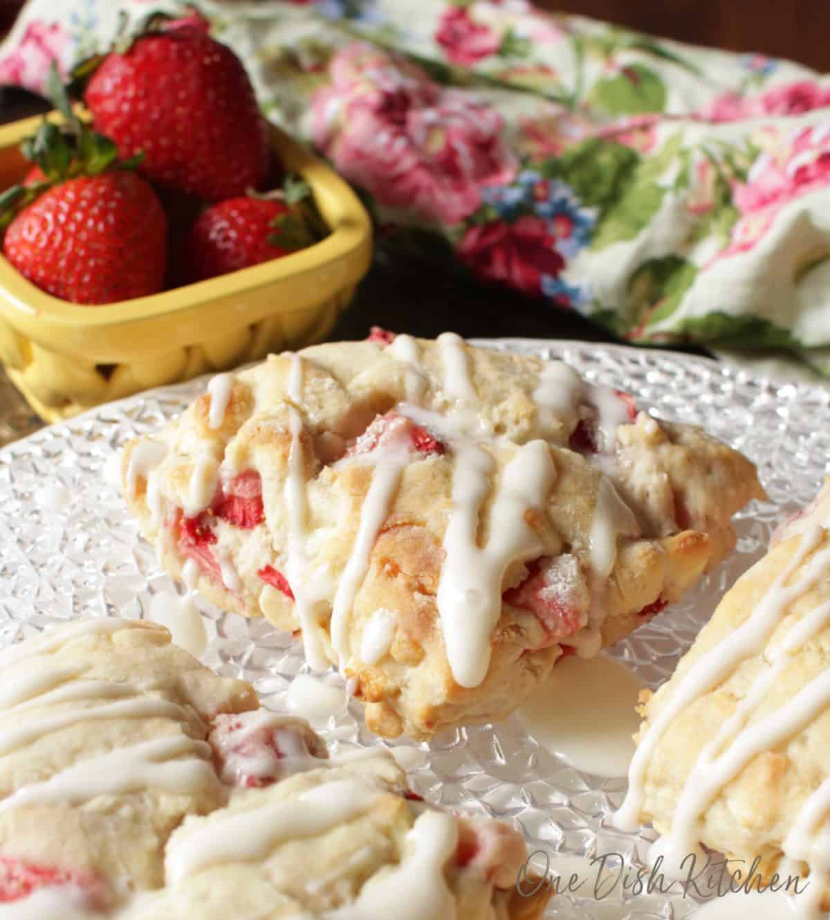 A closeup of strawberry scones with melted white chocolate drizzled over the top plated next to a small bowl of strawberries and a floral cloth napkin
