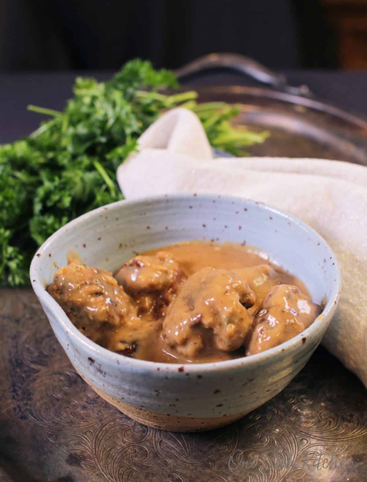 A small bowl of Swedish meatballs on a metal tray next to a bunch of parsley and a white cloth napkin.